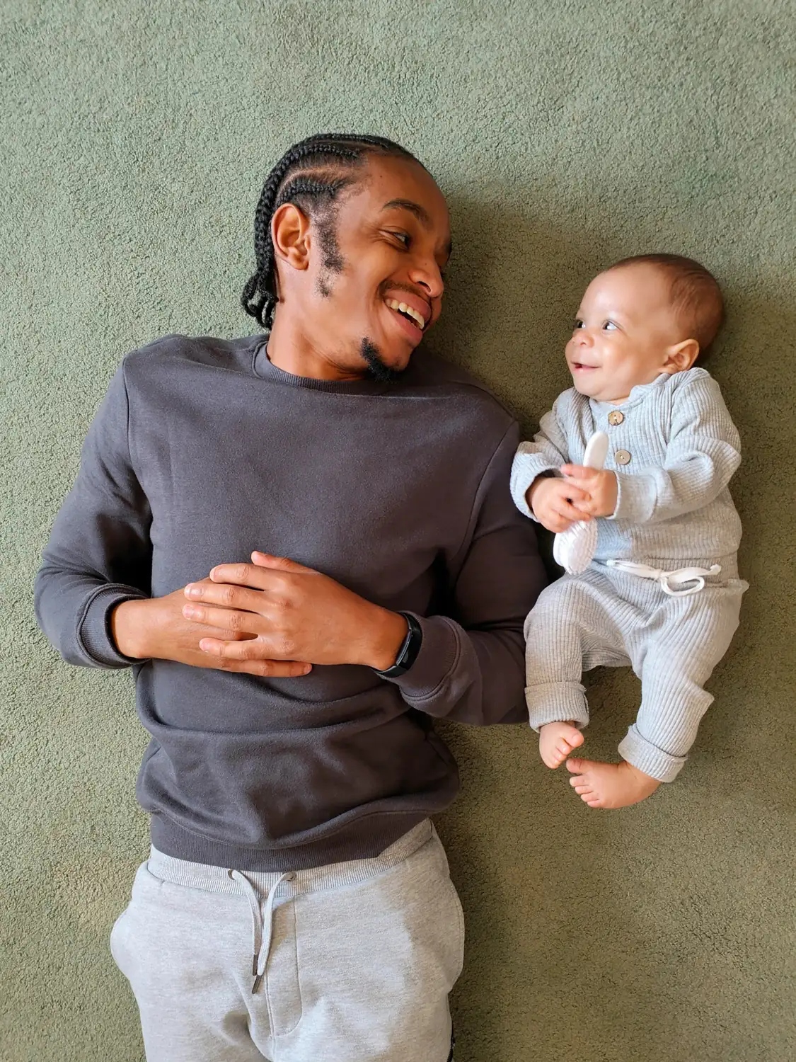 A man and a baby lying side by side on the carpet, grinning at each other