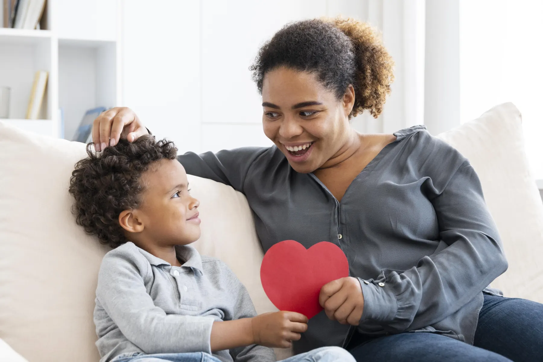 A carer and a young boy smiling at each other on the sofa, holding a paper heart