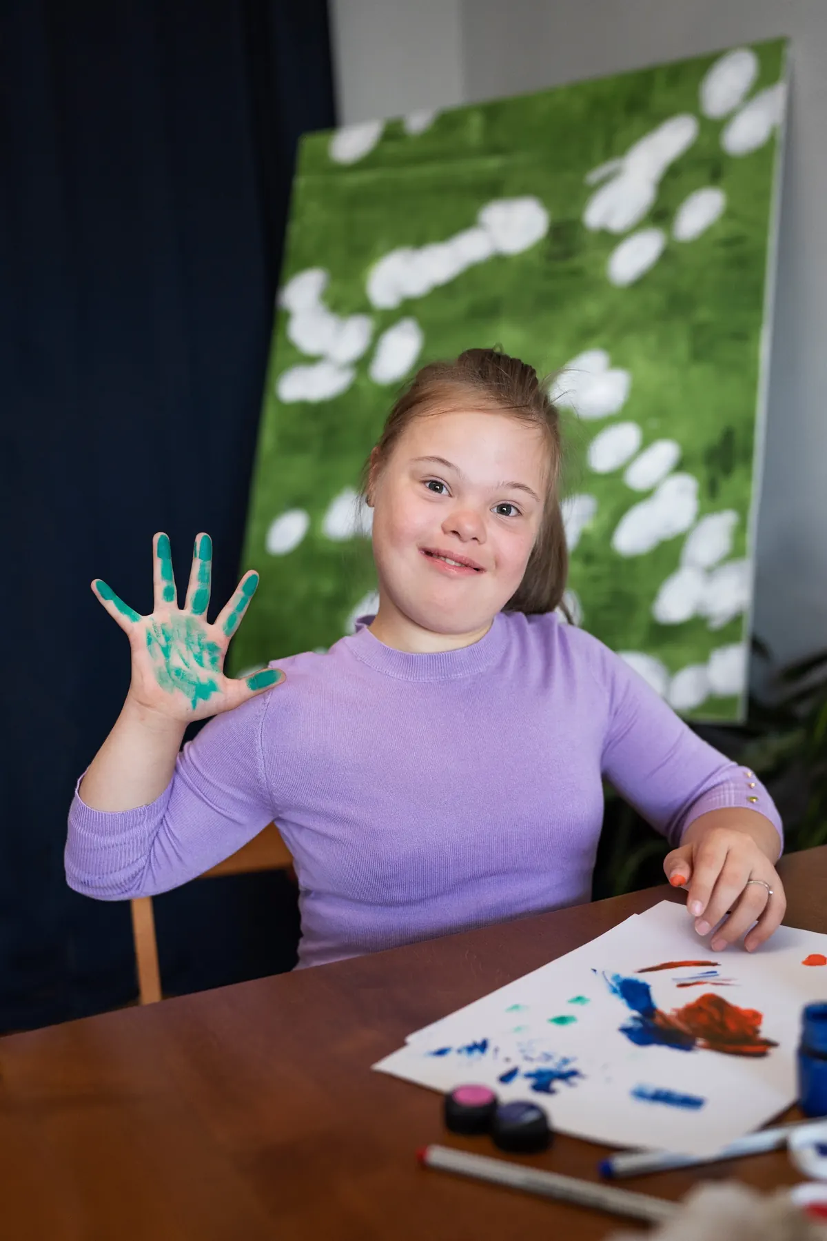 A girl at the table proudly showing a hand covered in paint